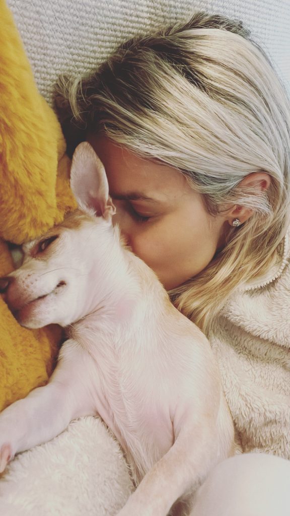 A woman resting her head against a small white dog on a couch